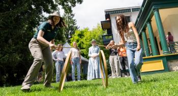 A Parks Canada interpreter plays hoop and stick with a student outside of Bellevue House while visitors observe alongside a guide in historic costume.