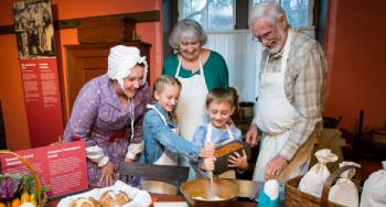 Seniors and children participate in an activity in the kitchen at Bellevue House National Historic Site while a guide in historic costume observes.