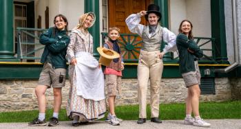 Parks Canada interpreters in uniform and in historic costume in front of Bellevue House with a youth wearing a costume vest and holding a straw hat.