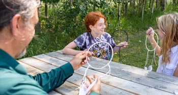 2 kids and 1 Parks Canada employee making dreamcatchers on a picnic table in nature